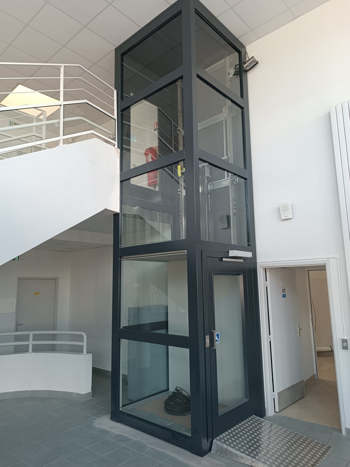 Élévateur pour personnes à mobilité réduite à gaine vitrée installé à côté de l’escalier du collège Les 7 Épis